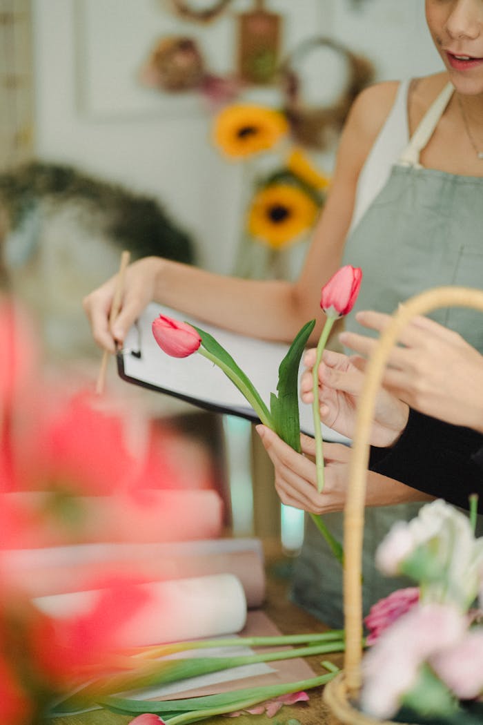 services-img-04 Unrecognizable female coworkers standing at table with tulips and writing notes on clipboard while working in florist atelier with blurred background